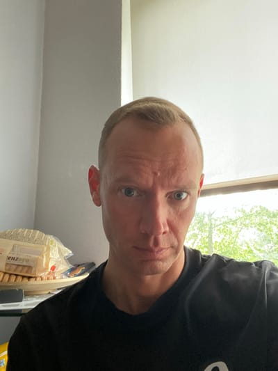 This media file is a selfie taken by a person in Tartu, Estonia. The person is a fair-skinned adult with short, light brown hair and light blue eyes. They are wearing a black t-shirt. The selfie is taken from a low angle, looking up at the person's face, which is slightly tilted. The person's expression appears neutral.
The background suggests an indoor setting near a window. Light filters in from the window, illuminating the scene and casting subtle shadows. Outside the window, green foliage is visible, indicating that it is daytime and likely spring or summer. Part of a white wall or ceiling is visible in the foreground. To the right of the person, a package of what appear to be cookies or biscuits is visible, with some text on the packaging. The text is partially obscured and difficult to read clearly, but "enzovo" is discernible. The overall lighting suggests it is daytime.
