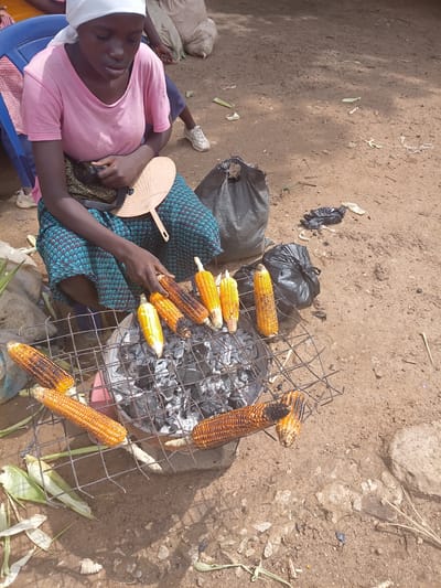A young woman, wearing a white head covering and a pink t-shirt, sits on a blue plastic chair outdoors, in what appears to be a marketplace in Tilde, Nigeria. She is grilling corn over a charcoal brazier. Her mid-section is adorned with a patterned cloth. In her right hand, she holds a wooden fan, possibly for fanning the coals. Several ears of corn are arranged on a wire rack placed over glowing embers. Some of the corn is already roasted, with charred kernels visible. In the background, there are bags, including a large black one, and discarded items on the dusty ground. The overall environment suggests a bustling, outdoor vendor setting.