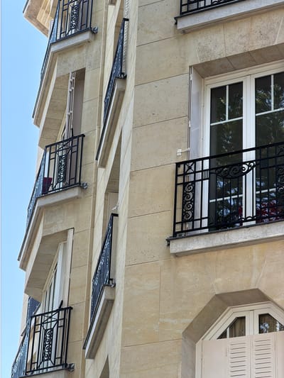 This is a low-angle, eye-level, close-up shot of the facade of a building, possibly an apartment building, located in Neuilly-sur-Seine, France. The building is constructed of light beige stone blocks. Several windows and balconies are visible, all with black wrought-iron railings featuring decorative scrollwork and vertical bars. Some windows have white frames and white louvred shutters. The sky, a clear, light blue, is visible in the upper left portion of the image. The light suggests it is daytime.