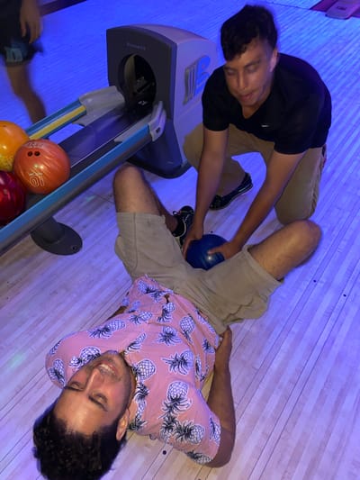 A person in a pink shirt with a pineapple pattern lies on their back on a bowling alley lane. Their legs are splayed and they are smiling with their eyes closed. Another person in a black shirt and tan shorts is kneeling next to them, holding a blue bowling ball. The bowling ball is positioned between the kneeling person's hands and the lying person's legs. In the background, a Brunswick bowling ball return machine is visible, with orange and red bowling balls in the rack. The bowling alley is lit with blue lights. The location is the city of Waynesboro, United States of America.