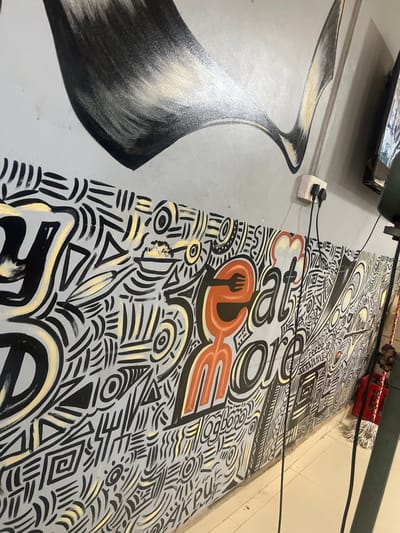 This image captures an indoor scene in Barako, Nigeria, showcasing a vibrant wall mural with an abstract and doodle-like aesthetic. The mural is a prominent feature, covering a large section of the wall with intricate patterns in black, light grey, and gold/yellow hues, incorporating geometric shapes such as circles, triangles, spirals, wavy lines, and zigzags.
A key element of the mural is stylized orange text, which clearly spells out "ORDER," with the letter 'E' creatively integrated with a black fork icon, strongly suggesting a food-related theme. Other orange letters are partially visible, reinforcing a culinary context.
To the left of the mural, the wall is plain light grey, adorned with a separate abstract, flowing design in black, white, and gold. An electrical outlet, white with two black cables plugged in, is mounted on this grey section, with the cables running across the vibrant mural.
In the upper-left corner, a dark, reflective object, possibly part of a fixture or shelf, is partially visible. Towards the right side of the image, a green pipe or beam stretches across the top. Below this, a red plastic crate containing objects (possibly bottles) and a white mop head are visible on a light beige tiled floor, indicating a commercial or service environment such as a restaurant or cafe. The overall lighting suggests an indoor setting.