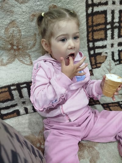 A young girl with blonde hair tied in two ponytails is sitting on a patterned blanket, eating ice cream. She is wearing a pink tracksuit with ruffled sleeves. The girl has her fingers in her mouth, and she is holding an ice cream cone in her right hand. The blanket has a beige background with brown floral and geometric patterns. The image appears to have been taken indoors, likely in a residential setting in Krasny Gulyai, Russia.