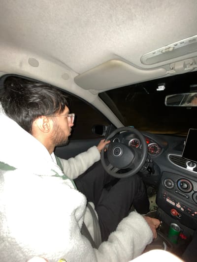 A man sits in the driver's seat of a car, wearing glasses and a fluffy white jacket. He has dark hair and a beard. He is looking out the window, holding the steering wheel. The car's interior is visible, including the dashboard, rearview mirror, and sun visor. It appears to be nighttime. The image is taken at an odd angle, as if the photographer is lying on their side.