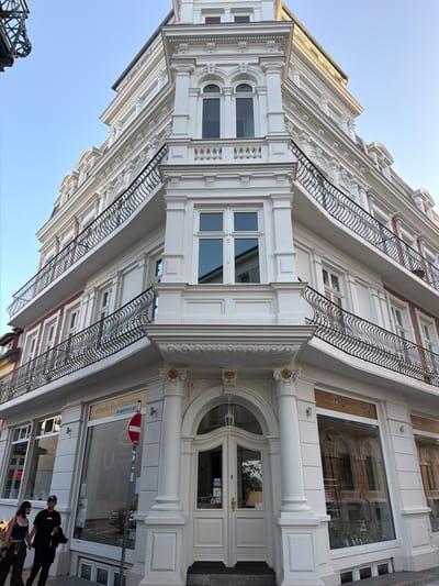 The image shows a white, ornate building with multiple stories and balconies. The building has a corner entrance with a large arched doorway and double doors. The windows are tall and rectangular, with decorative frames. There are black wrought-iron railings on the balconies.
In front of the building, two young adults are walking. A young woman on the left is wearing a dark blue tank top and dark pants. A young man on the right is wearing a black t-shirt and dark pants, holding a jacket. They are walking hand-in-hand.
To the left of the entrance, there is a shop window with a sign that reads "stilvolle Einrichtungen Alexandrinenstraße". A red and white circular stop sign is mounted on a pole in front of the shop window. The street appears to be paved with stone. The sky is clear and blue.
This building is located at Alexandrinenstraße in Rostock, Germany.