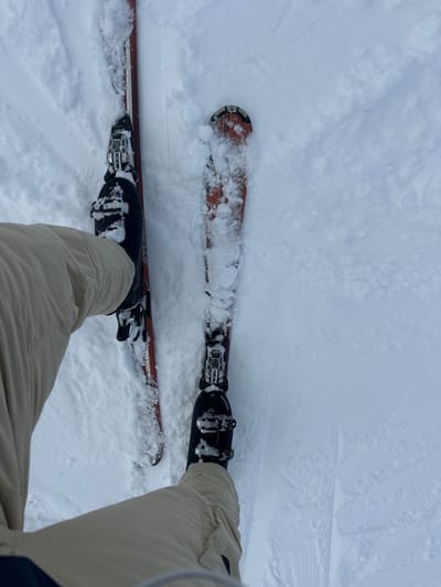 A first-person view depicts the lower body of an individual standing on skis in deep, fresh snow within Bad Hofgastein, Austria. The person wears light beige or tan ski pants and dark ski boots, which are secured into ski bindings on two orange-red skis. The skis are partially submerged in undisturbed white snow, with some visible snow accumulation on their top surfaces and bindings. The immediate environment consists entirely of snow, showing faint depressions or previous tracks around the skis.