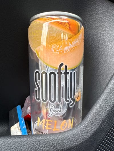 A "SOOFTY Drink MELON" flavored aluminum beverage can is positioned upright within a vehicle's interior, likely in a cup holder or door pocket. The can's label features prominent images of sliced cantaloupe melon against a translucent background, with the brand name "SOOFTY" in black outlined text, and "Drink" and "MELON" in orange lettering beneath. "NATURAL AROMA" is partially visible at the bottom of the label. The can is sealed, appearing full. To its immediate left, partially obscured, is minor debris, including a blue item and crumpled translucent material. The immediate environment consists of dark, textured vehicle interior surfaces; a smoother black material on the left and a coarser, grey-black textured material on the right. The scene is located in Kaysersberg Vignoble, France.