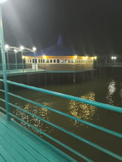 The image depicts a wooden restaurant building situated on stilts over a body of water, likely at night. The building has a distinctive conical blue roof and wooden exterior walls. Numerous windows line the sides of the structure, illuminated from within. A series of lights are strung along a walkway leading to the building, and more lights are visible further in the background, suggesting a pier or boardwalk. The water reflects the lights, creating shimmering patterns on its surface. The foreground features a teal-colored railing and a wooden deck, also teal. The scene appears to be a waterfront dining establishment, possibly a tourist attraction. The location is identified as Bayet, Kyrgyzstan. There are no people visible in the image.