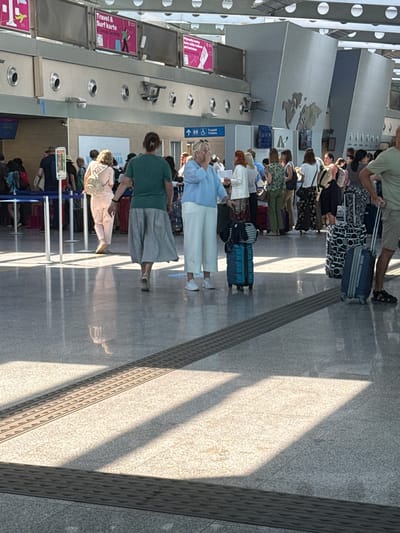 The image depicts a queue of people at what appears to be an airport or train station in Golubovci, Montenegro. The setting is indoors with a polished gray floor, columns, and a wall with what looks like a map of Europe. Sunlight is streaming in from the right, casting long shadows across the floor, suggesting it is daytime, likely afternoon.

The subjects are primarily people standing in line, some with luggage. They are dressed in casual clothing suitable for travel. Visible objects include various suitcases, some rolling, and stanchions with blue tape creating the queue. There are also promotional posters on the wall to the left, one of which reads "Travel & Surf Karte". Another sign appears to be a directional sign with icons for accessibility and toilets.

The activity is people waiting in line, presumably for check-in, boarding, or security. The mood appears neutral, as is typical for travel waiting areas. The lighting and shadows add a dynamic element to the scene. The overall impression is one of everyday travel activity.