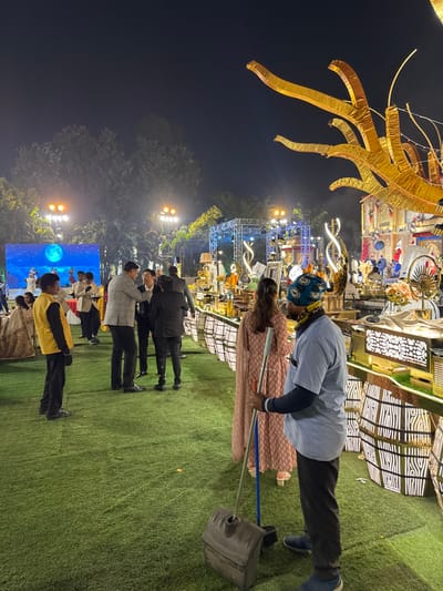 An outdoor event at night features a large golden structure. A man wearing a blue shirt and a patterned head covering sweeps green turf with a blue broom and a large black dustpan. Several people stand behind them, looking toward a lit stage with screens displaying images, possibly the moon. The area is illuminated by strung lights and stage lights.