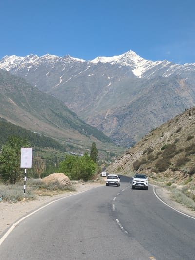 The image shows a scenic mountain landscape with a road curving through a valley. The road is paved and has white lane markings. Two white SUVs are driving on the road, one in the foreground and one further back. A white traffic sign with speed limit indicators of 50 and 40 is visible on the left side of the road. The surrounding environment consists of steep, rocky mountains with sparse vegetation and some snow-capped peaks in the distance. The sky is clear and blue. The image was taken in the vicinity of Gondhla, India.