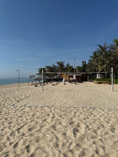 This is a bright, sunny, outdoor, eye-level, full shot of a sandy beach in Cam Lâm, Vietnam, with a volleyball net in the foreground. The beach has light brown sand with visible footprints. A blue line marks the boundary of the volleyball court. In the middle ground, there is a beach volleyball court with a net and poles. Behind the court, there are several thatched-roof huts, wooden tables, and chairs, suggesting a beachside dining or relaxation area. Palm trees are visible in the background, lining the edge of the beach. The ocean stretches out to the left, with gentle waves lapping the shore. A tall lamp post stands on the left side of the beach. In the distance, a structure resembling a wooden ship or boat is visible, adorned with a flag. The sky is a clear, bright blue. There are no people actively playing volleyball or in the immediate vicinity of the court.