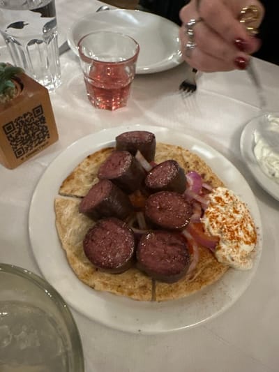 The image captures a dining table scene, likely indoors, in Agios Dimitrios, Greece. The primary subject is a white plate holding a meal, consisting of several thick slices of dark, cooked sausage (possibly blood sausage or a similar specialty) resting on pieces of pita bread. The sausage and pita are topped with thinly sliced red onions and a generous dollop of white, creamy sauce, which is sprinkled with red paprika.

To the left, a person's hand with red nail polish and multiple rings is visible, holding a silver fork, suggesting an ongoing or imminent meal. Another small white dish containing more of the white creamy sauce is partially visible in the upper background.

On the white tablecloth, a clear glass with a pinkish hue is lying on its side, while another clear glass is partially visible at the bottom right. A small, light-colored wooden block with a QR code printed on it is also seen in the foreground, with the partially visible text "CONNECT WIFI" beside the code. The lighting suggests an evening or indoor setting.