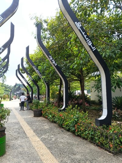 The image displays a paved outdoor walkway within the city of Sarandi de Cuadra, Uruguay, under bright daylight. A series of arching, black and white decorative structures, each bearing white Arabic script such as "الودود" (Al-Wadud) and "الواسع" (Al-Wasi'), lines the right side of the path. The environment is rich with green foliage from numerous trees and shrubs, which feature small orange-red blossoms. A low, dark metal fence separates the shrubbery from the walkway in the foreground. The walkway itself is composed of gray, speckled concrete, incorporating a central strip of yellow tactile paving. In the middle ground, three adult males are visible standing on the walkway, facing away from the camera. One male wears a white t-shirt and dark pants, while another wears a black t-shirt and light-colored pants. Black planters with green plants are placed along the walkway, and a portion of a green cylindrical container is visible on the far left. A white building or structure is partially visible through the trees in the distant background on the right.
