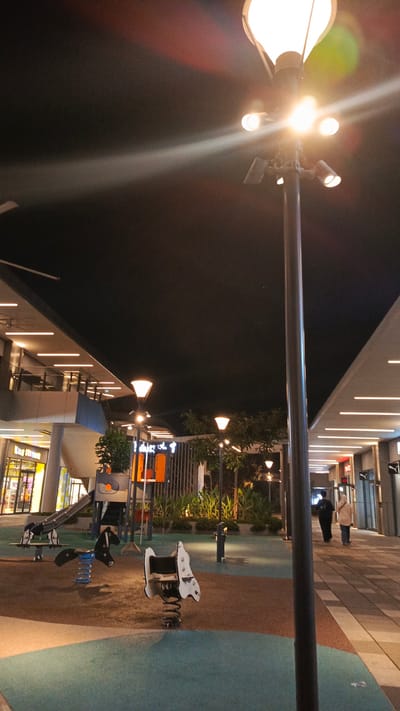 The image depicts a modern outdoor playground area at night, illuminated by overhead streetlights and architectural lighting. The playground features several spring riders, including two horse-shaped ones, a slide, and a climbing structure. The ground is a mix of light brown and teal-colored soft surfacing. To the left, a building with multiple levels and illuminated window fronts is visible. The right side of the image shows a paved walkway with two individuals walking away from the camera. The sky is dark, indicating nighttime. This scene is located in the city of Wanasari, Indonesia.