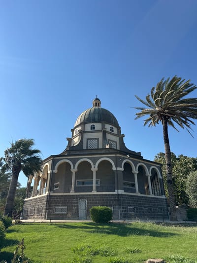 The image shows the Church of the Beatitudes, a Roman Catholic church located on a hill overlooking the Sea of Galilee in the Emek HaYarden Regional Council, Israel. The church has a distinctive octagonal shape with a central dome topped by a cross. The lower level of the church features a colonnade with rounded arches, constructed from dark stone. Above the arches, the building is made of lighter colored stone, with decorative window grates and arched windows. Two palm trees are visible, one tall and slender to the right of the church, and another shorter, bushier one to the left. The foreground is a well-maintained green lawn, with some smaller plants and shadows cast by the trees and the church. The sky is a clear, bright blue, suggesting a sunny day. The scene is peaceful and serene, with no people or animals visible.