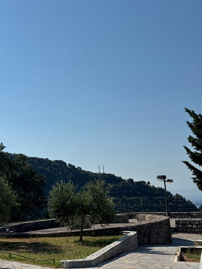 The image shows a view from a stone-paved area with some vegetation. In the foreground, there is grass and a low stone wall. Several trees are visible, including olive trees. A curved stone wall with steps leads to a higher level. A modern street lamp with two lights stands on the right side of the paved area. In the background, a densely forested hill rises, and in the distance, the sea can be seen. The sky is clear and blue. Two telecommunication towers are visible on top of the hill. The scene is an outdoor environment, likely a park or viewpoint.