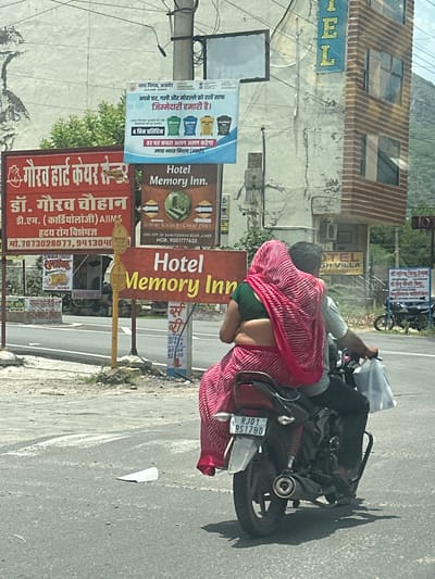 This is a medium shot, taken from a vehicle, of a couple riding on a motorcycle in Ajmer, India. The couple is dressed in traditional Indian clothing, with the woman wearing a pink and white striped sari. The motorcycle has a license plate that reads "RJ01 BS1780". In the background, there are several signs, including one for "Hotel Memory Inn." There is also a building with "HOTEL" written on it vertically in large blue letters. The environment appears to be a city street on a sunny day, with some vegetation and mountains visible in the distance.