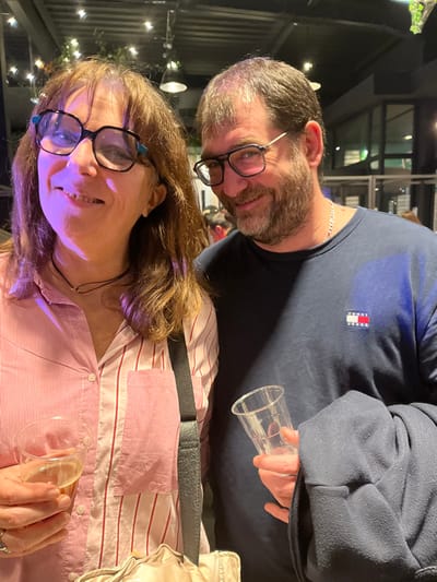 A couple poses close together. The woman wears a striped pink shirt and glasses, smiling toward the camera while holding a glass with a yellowish liquid. The man behind her has a beard and glasses. He wears a dark blue shirt with a logo and holds an empty plastic cup. They appear to be inside a building with string lights in the background. A black strap is visible across the woman's torso.