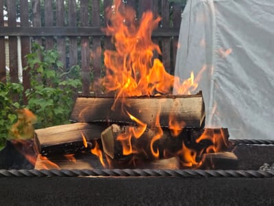 A bonfire is burning in a metal grill in a backyard setting. The fire is contained within a dark-colored grill with a twisted metal grate. Several logs of wood are stacked inside the grill, with bright orange flames consuming them. Some of the wood is charred black from the heat. In the background, a wooden fence runs horizontally across the frame, with green foliage visible behind it. To the right of the fire, a large white tarp or cover is partially visible, also with flames flickering nearby. The scene appears to be set outdoors during the daytime, possibly in the evening given the intensity of the fire. There are no people visible in the image, and no text is present. The overall impression is of a warm and active outdoor scene.