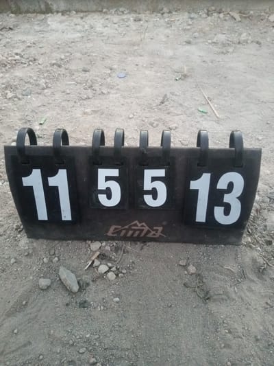 The image shows a scoreboard on a dirt and gravel surface. The scoreboard is black with flip numbers displaying "11", "5", "5", and "13". There are four black metal loops at the top of the scoreboard, each holding a flip number panel. A logo appears on the lower front of the scoreboard. The surrounding environment is an outdoor area with uneven, sandy ground containing small stones and debris. There are no people or animals visible in the image.