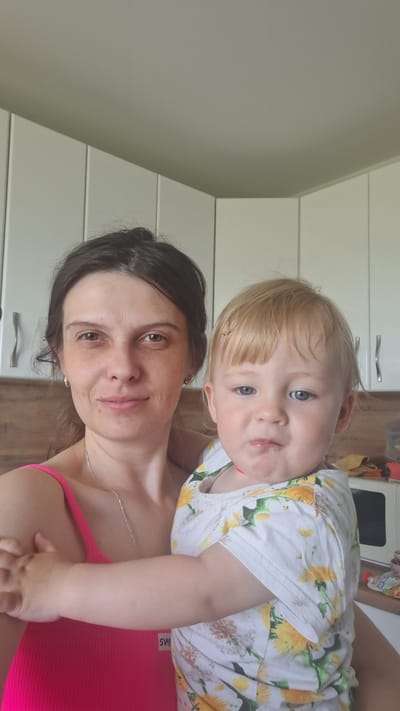 A medium shot shows a woman holding a toddler in a kitchen. The woman, with dark hair pulled back, is wearing a bright pink tank top and a silver necklace. She is looking at the camera with a slight smile. The toddler, with blonde hair and blue eyes, is wearing a white shirt with a yellow floral pattern. The toddler's face is turned slightly away from the camera, and they appear to be making a facial expression. The background consists of white kitchen cabinets and a wooden countertop. The scene is illuminated by natural light. The location is identified as the city of Myza (Inshinskii S/O), Russia.