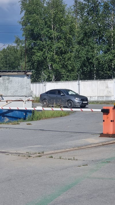 The image depicts an exterior scene in the city of Megion, Russia, featuring a gray sedan parked behind a white and red striped barrier gate. The car has its headlights on, and its license plate is visible. The environment includes a concrete building to the left, a white fence in the background, and lush green trees. The ground is paved with asphalt, and there is a drain grate on the pavement. An orange barrier gate mechanism is visible on the right side of the image. The sky is partly cloudy.