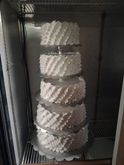 A five-tiered cake decorated with white frosting ruffles is displayed inside a commercial refrigerator. The cake layers are stacked on silver scalloped cake stands. The refrigerator has a textured metallic interior and wire shelves. The location is in the city of Guldala, Kazakhstan.