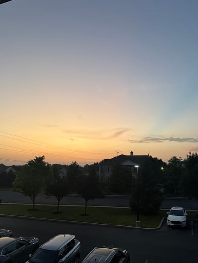 The image depicts an outdoor scene at dusk in Allen Township, United States of America. The sky displays a gradient of colors, transitioning from a pale orange at the horizon to a soft blue at the top, with wispy clouds scattered across the expanse. Several power lines are visible traversing the sky horizontally.

In the middle ground, a building with a pitched roof and a prominent chimney is partially obscured by trees. Streetlights are illuminated in front of the building. A row of trees lines the edge of a paved area, which appears to be a parking lot.

The foreground shows a section of the parking lot with several vehicles parked. These include a white SUV, a silver SUV, and a dark-colored sedan. The overall impression is a quiet residential or commercial area at the end of the day.