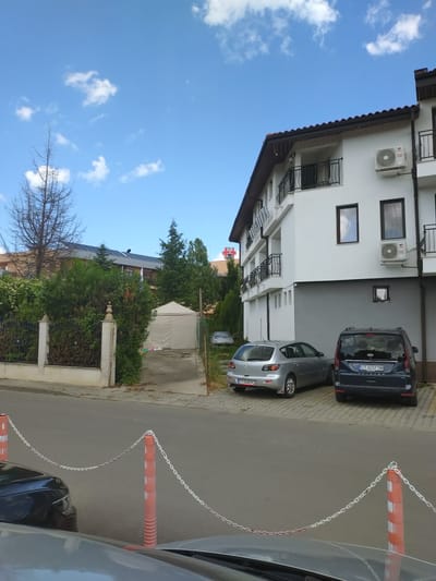 The image captures a street scene in Yagoda, Bulgaria, with a modern building and parked cars. In the foreground, a grey car and a dark blue minivan are parked on a paved area. The minivan has the license plate "CT 3237 TM". To the left of the minivan is a silver hatchback with the license plate "CT 0540 PC". The street is bordered by orange traffic cones connected by a white chain.
Behind the cars, a white building with multiple balconies and air conditioning units is visible. The building has a brown tiled roof. Adjacent to the building, a green hedge runs along a white fence. Further back, there's a white tent structure. A cross symbol is visible on a building in the distance. The sky is blue with scattered white clouds. The environment appears to be a residential or commercial area with trees and vegetation.