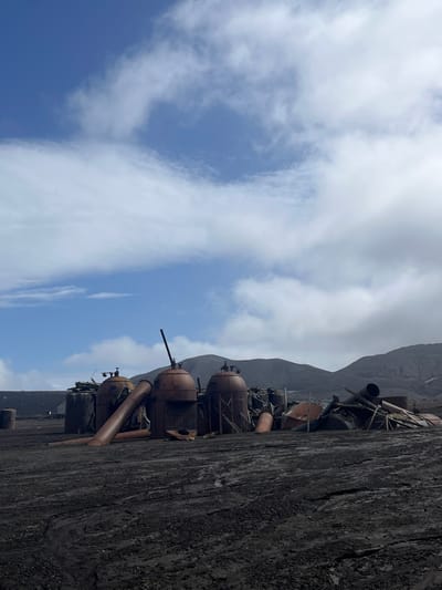 Rusted, cylindrical industrial components with pipes and metal supports are stacked against a textured, dark gray or black surface. A mountain rises in the background, partially obscured by clouds. The sky is a mix of blue and white, with puffy clouds scattered across the upper portion of the image. The viewpoint is from below, looking upwards at a slight angle.