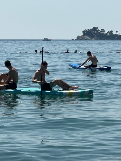 In the city of Đurovići, Montenegro, on a bright, sunny day, several individuals are enjoying water activities. In the foreground, two young men are seated on separate paddleboards. The man on the left, wearing dark shorts, is facing away from the camera. The man on the right, also in dark shorts, is holding a paddle and facing left. Behind them, several other individuals are visible in the water, with only their heads above the surface. In the background, a small boat is seen on the horizon, and a rocky island covered in trees is to the right. The sea is calm with gentle ripples, and the sky is clear.