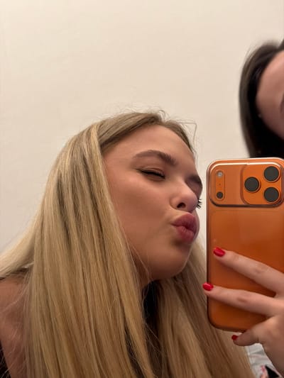 A woman with blonde hair purses her lips, partially obscured by an orange smartphone held up with a hand with red nail polish. The phone's camera array is visible. Reflected in the phone's surface is a darker-haired individual, partially cut off at the top, creating an upside-down mirror effect. The background is a plain, light-colored wall. The image is taken from a slightly tilted, low-angle perspective.