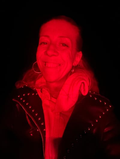 The image shows a medium close-up, self-portrait of a woman with auburn hair, wearing a black leather jacket adorned with studs, and a red scarf or neck warmer. The lighting is predominantly red, casting a strong red hue over the entire scene. The woman has a wide smile, revealing her teeth. She is wearing large hoop earrings. The background is entirely black. The image was taken at an unknown location in the city of Krautscheid, Germany.