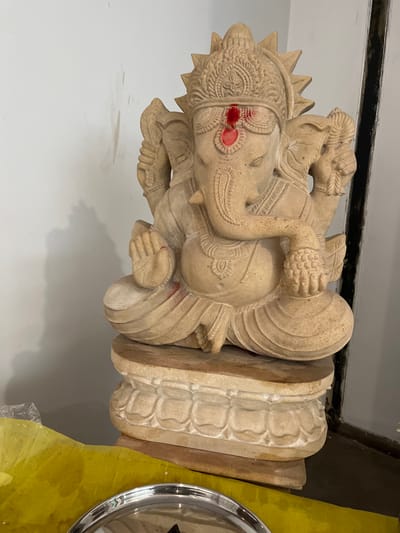 A tan-colored, carved stone statue of the Hindu deity Ganesha sits on a pedestal, facing forward. The statue is adorned with a red tilak on its forehead and red markings on its trunk and hands. Ganesha is depicted in a seated position, with its trunk curving to the left and its hands held in devotional gestures. The pedestal features carved lotus petal motifs. To the left of the statue, a bright yellow cloth is visible, partially covered by plastic wrap. The background consists of a white wall with some smudges and a dark vertical element to the right. The image appears to be taken indoors, possibly in a residential or temple setting in Greater Noida, India.