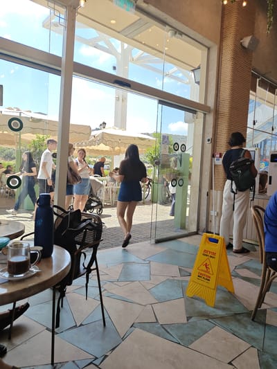 The image depicts a bright, sunny day outside what appears to be a cafe or restaurant. The scene is viewed from inside, looking out through a large glass door and window. Several people are visible in the outdoor area and entering the establishment.
In the foreground, a round table with a coffee cup and a water bottle is prominent. In the background, patrons are seen mingling and walking. One woman in a denim skirt is walking away from the camera, while another woman wearing glasses and a white top is standing and talking. A man in a white t-shirt and shorts is also visible, along with a stroller and a woman pushing it. Further in the background, several cafe umbrellas provide shade.
A prominent yellow caution sign with a wet floor symbol and text in Hebrew, English, and Arabic is placed on the tiled floor near the entrance. The text "CAUTION WET FLOOR" is clearly legible, along with Hebrew text "זהירות רצפה רטובה" and Arabic text. The overall atmosphere appears casual and relaxed, suggesting a typical daytime scene. The bright sunlight and clear sky indicate good weather. The setting is likely an outdoor seating area of a cafe, perhaps in a town square or promenade, given the pedestrian traffic and the presence of umbrellas.