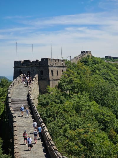 The image depicts a section of the Great Wall of China, specifically in the Huairou District of China. The wall, constructed of grey stone, snakes along a lush green, tree-covered hillside under a bright blue sky with scattered white clouds. Several stone watchtowers with crenellated battlements are visible along the wall. A group of tourists, diverse in age and attire, are seen walking up a steep stone staircase on the wall. They are spaced out and moving in the same direction. The environment is natural, with dense foliage and a clear sky.