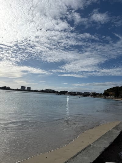 The image displays a wide-angle view of Anse Vata Bay in Noumea, New Caledonia, France, under clear daylight conditions. The sky occupies the upper half of the frame, exhibiting a vibrant blue color with extensive coverage of white, textured altocumulus clouds, most dense towards the upper left where the sun's brilliance is inferred.
The central and lower portions of the image are dominated by the calm, light blue-grey waters of the bay. A discernible vertical streak of sunlight reflects on the water in the middle-right. A line of dark buoys extends horizontally across the bay from the left shore, segmenting a portion of the water. Two small, indistinct human figures are visible within the bay's waters, positioned near the reflected sunlight.
The immediate foreground on the bottom right reveals a narrow strip of light-colored sand, upon which two human footprints are faintly impressed. This sand meets a concrete or paved retaining structure that forms the extreme right edge of the scene.
The distant shoreline across the bay, spanning from left to center-right, features urban development. On the left, several multi-story buildings, including two more prominent high-rise structures, are visible. Towards the center-right, a long pier or similar structure extends into the bay, connecting to additional buildings along the distant shore. A vegetated hillside rises on the far right of the background.