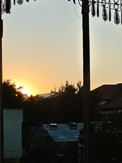 The image depicts a sunset over a residential area in Mława, Poland. The sun, a bright orb of orange and yellow, is partially obscured by the silhouettes of trees and buildings, casting a warm glow across the sky. The sky transitions from a soft yellow near the horizon to a pale blue at the top.
In the foreground, a dark, vertical utility pole stands prominently, with decorative items hanging from a wire strung across its top. The items appear to be made of dark, patterned material, possibly fabric or lace, with a looped design. A thin, coiled wire hangs loosely from the pole, suggesting electrical or communication infrastructure.
Behind the pole, a dense layer of dark green trees forms a natural barrier, with the setting sun peeking through their branches. Above the trees, rooftops of houses are visible, with one building displaying a dark red tiled roof. Further into the scene, a greenhouse-like structure with a corrugated metal roof and several skylights is partially visible amidst the foliage. To the left of the scene, a light-colored rectangular building stands, its upper portion cut off by the frame.
The overall mood of the image is tranquil and serene, capturing the peaceful end of a day in Mława. The warm light of the sunset and the quiet residential setting evoke a sense of calm. No visible text is present in the image.