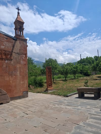 The image depicts a view of a stone structure, possibly a church or monastery, with a bell tower adorned with a cross. The structure is made of reddish-orange stone, and features intricate carvings on its facade. Adjacent to the building is a paved walkway made of large, grey stone slabs, with some grass and dry vegetation visible in the background.

Beyond the immediate structures, the scene opens up to a lush green landscape with numerous trees. In the distance, rolling hills and mountains can be seen, partially shrouded in a light haze. The sky is a vibrant blue, dotted with fluffy white clouds. Two telephone poles are visible against the sky, suggesting the presence of modern infrastructure in the otherwise natural setting.

A stone khachkar, an Armenian cross-stone, stands upright in the middle ground, carved with intricate patterns. To the right of the khachkar, a stone bench or seating area is visible. The lighting suggests it is daytime, likely late morning or early afternoon, with bright sunlight casting shadows on the ground. The overall impression is one of a serene, historical site located in a scenic, rural area of Armenia. No people are visible in the image. There is no discernible text in the image.