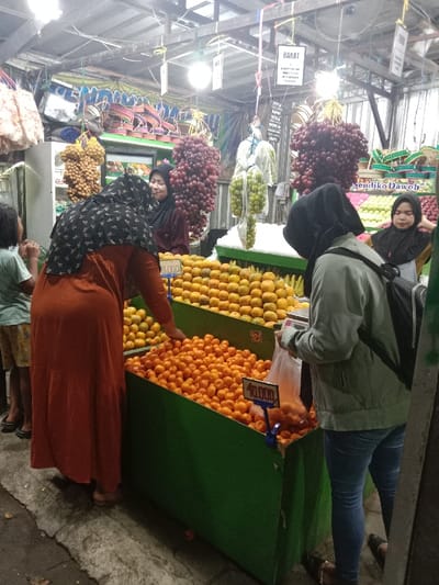 An indoor market stall in Sekopek Wetan, Indonesia, displays a variety of fresh fruit under artificial lighting. Four women and one child are present. In the foreground, a woman wearing an orange long dress and a dark patterned headscarf is bent over a green display bin filled with numerous small oranges, her right hand reaching into the fruit. To her left, a young child in a light shirt and patterned shorts stands beside her. To the right, another woman wearing a light green jacket, blue jeans, a black headscarf, and carrying a black backpack, stands facing a similar green fruit bin, holding a clear plastic bag in her left hand.
Behind the fruit display, two women, likely vendors, stand by the counter. The woman on the left, wearing a dark red top and a dark headscarf, smiles and observes the customer in orange. The woman on the far right, wearing a black headscarf, is partially visible.
Overhead, several clusters of dark red grapes, light brown oval-shaped fruit (likely longan), and green fruit in plastic bags hang from the ceiling structure. Multiple bare lightbulbs illuminate the scene. Two green display bins are filled with oranges, one with a visible price tag reading "10.000". Another price tag, partially obscured, is visible on the upper bin. Various signs with Indonesian text, including "BAKAT" and "SENDIKO DAWOH", are visible on the upper structure. The floor is an unadorned concrete surface.