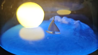 A small, white toy sailboat with a single sail floats on a shimmering blue surface that resembles water. The water has white, foamy crests, suggesting movement or waves. To the left of the sailboat, a large, bright yellow orb, likely a lamp or light source, illuminates the scene and casts a soft glow onto the blue surface. In the background, a pale, oval-shaped object is visible, possibly another light or a reflection. The overall impression is a miniature, stylized seascape. The image does not contain any identifiable elements or context to link it to the city of Шемордан, Russia.
