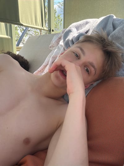 The media file is a close-up, low-angle selfie taken outdoors. The primary subject is a young person with light brown hair, wearing a gray towel draped over their head and shoulder. They are lying down, with their right hand covering their mouth and their gaze directed towards the camera. Their chest and upper torso are visible, and they appear to be shirtless.

The background suggests an outdoor setting, possibly a patio or lounge area. Green fabric elements, likely from outdoor furniture like lounge chairs, are visible on the left side of the frame. Further back, trees and a bright blue sky with some clouds indicate it is daytime, likely sunny. An orange and gray patterned fabric, possibly a cushion or blanket, is visible behind the subject's head and to the right.

The overall mood appears relaxed and casual. The subject's hand gesture and direct eye contact suggest a playful or contemplative pose. The lighting indicates it is daytime, with sunlight illuminating the scene. There is no visible text in the media file. The provided location context of Khotianivka, Ukraine, suggests the geographical setting.
