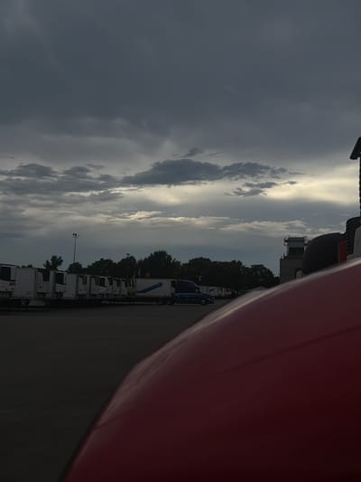 The image depicts a parking lot with several semi-trailer trucks and a building in the background under a cloudy sky. The foreground is dominated by the red hood of a vehicle. In the middle ground, a row of refrigerated trailers is parked, some bearing the brand "MARK." A blue semi-trailer truck is also visible, parked to the left of the trailers. The background features a line of trees and a multi-story building with industrial-looking structures. The sky is filled with dark, dramatic clouds, with a sliver of sunlight breaking through on the right side. The scene suggests an overcast or approaching storm. Based on the metadata, the location is Murfreesboro, United States of America.