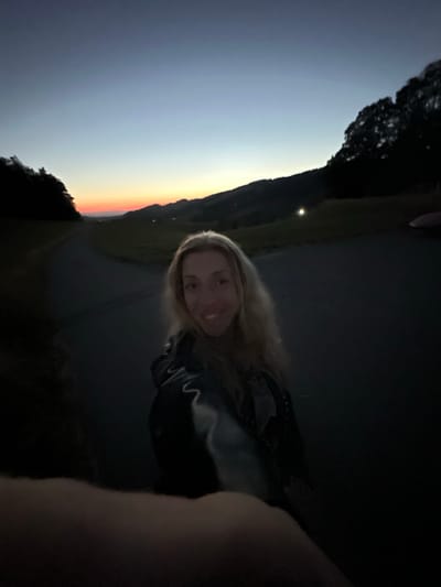 In St. Peter, Germany, a woman with blonde hair is pictured taking a selfie at dusk. She is wearing a dark jacket. The background shows a road stretching into the distance, with hills and trees silhouetted against a twilight sky. The sky transitions from a pale blue at the top to orange and pink hues near the horizon. The lighting is dim, characteristic of sunset.