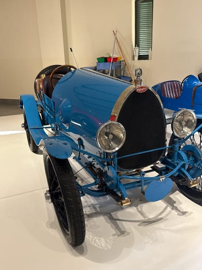The image shows a blue vintage Bugatti car, viewed from the front and slightly above. The car is indoors, likely in a museum or showroom, as evidenced by the plain white walls and artificial lighting. The car's front wheels, radiator, headlights, and part of the driver's seat are visible. The radiator has the Bugatti emblem. Details like the spoked wheels and the complex suspension system are also apparent. The location is likely a museum in the Stellenbosch Local Municipality, South Africa, based on the image prompt. There are no people visible in the frame.
