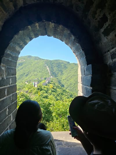 This media file captures a view through an arched opening in a stone wall, likely part of the Great Wall of China in Huairou District. The opening frames a lush, green mountainside covered in dense vegetation. A section of the Great Wall is visible winding along the contours of the mountain in the distance. The sky is a clear, bright blue, suggesting a sunny day, likely morning or afternoon due to the natural light.
In the foreground, two people are partially visible. One person, wearing a dark hat and a light-colored top, is holding a smartphone and appears to be interacting with it. Their hands and part of their face are visible. Another person, wearing a dark green jacket, is looking downwards and is mostly obscured by shadow. Their head and shoulders are the most prominent features. The perspective suggests they are standing on or near the wall, looking out through the archway. The weathered texture of the stone wall is evident, with individual bricks clearly distinguishable. The overall scene evokes a sense of travel and exploration, with the individuals taking in the expansive natural scenery and historical landmark.