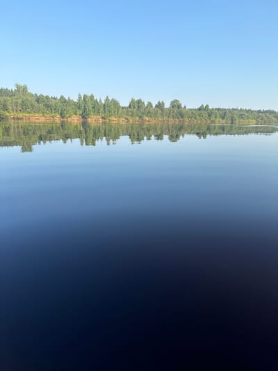 This image depicts a serene natural landscape, likely a body of water with a reflection of a treeline and a pathway. The dominant colors are deep blues and greens, with a clear, bright sky reflected in the water. The left side of the image is a gradient of dark to light blue, suggesting deep water or a transition in light. The right side features a dense row of green trees, with a sandy or dirt path running along the top of the treeline. The reflection in the water is nearly perfect, mirroring the trees and path with subtle ripples.

The scene appears to be set during daylight, with the bright sky indicating a sunny day. The stillness of the water suggests calm weather conditions. The location is identified as Slobodskoy, Russia, which conjures images of natural beauty typical of the region. There are no visible people or objects other than the natural elements. The overall mood is peaceful and tranquil, emphasizing the quiet beauty of nature. The reflection creates a sense of symmetry and depth. There is no visible text in the image.
