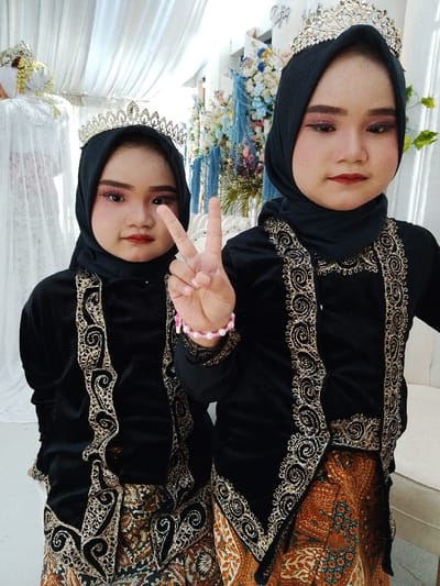 Two young females are centrally positioned in an indoor event setting in Juwana, Indonesia. Both wear black hijabs and gold-colored tiaras. Their attire consists of black velvet long-sleeved tops or jackets with elaborate gold embroidery, paired with patterned brown, orange, and white batik sarongs. Both individuals have visible makeup, including pink eyeshadow and red lipstick, and are looking directly at the camera. The female on the left, appearing younger, performs a V-sign with her right hand, which also displays a pink and white beaded bracelet. The background features white draped fabric, floral arrangements with blue, pink, and white elements, a partial white column with text, and a section of a white cushioned seat. A person in a white gown and an elaborate white headpiece is partially visible on the far left.