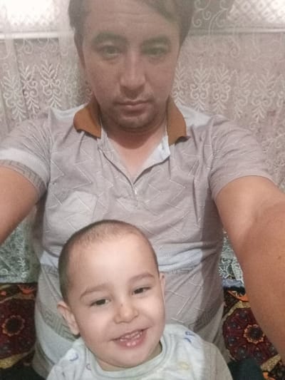A man poses with a toddler. The man, positioned behind the child, wears a short-sleeved shirt with a brown collar and a geometric pattern. The toddler, in the foreground, smiles, revealing his teeth. The backdrop consists of a patterned wall covering, and the lower portion reveals patterned fabric. The lighting appears somewhat dim, and the image seems to be a candid snapshot.
