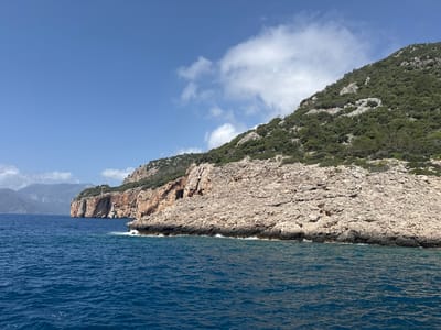 The image depicts a coastal scene near Kumluca, Turkey. A rocky headland extends into the deep blue sea. The headland features a base of light-colored, heavily eroded rock, topped with a dense layer of green vegetation. Further along the coast, another headland with similar rocky formations and sparse vegetation is visible in the distance. The sky is blue with scattered white clouds. Small waves are visible at the base of the headland where the water meets the rocks.